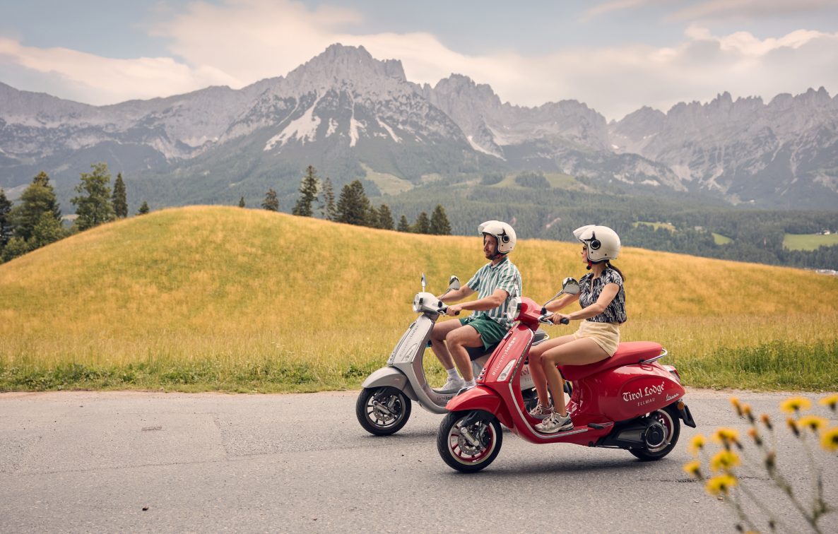 Zwei Personen fahren mit Rollern vor einer Bergkulisse bei Ellmau